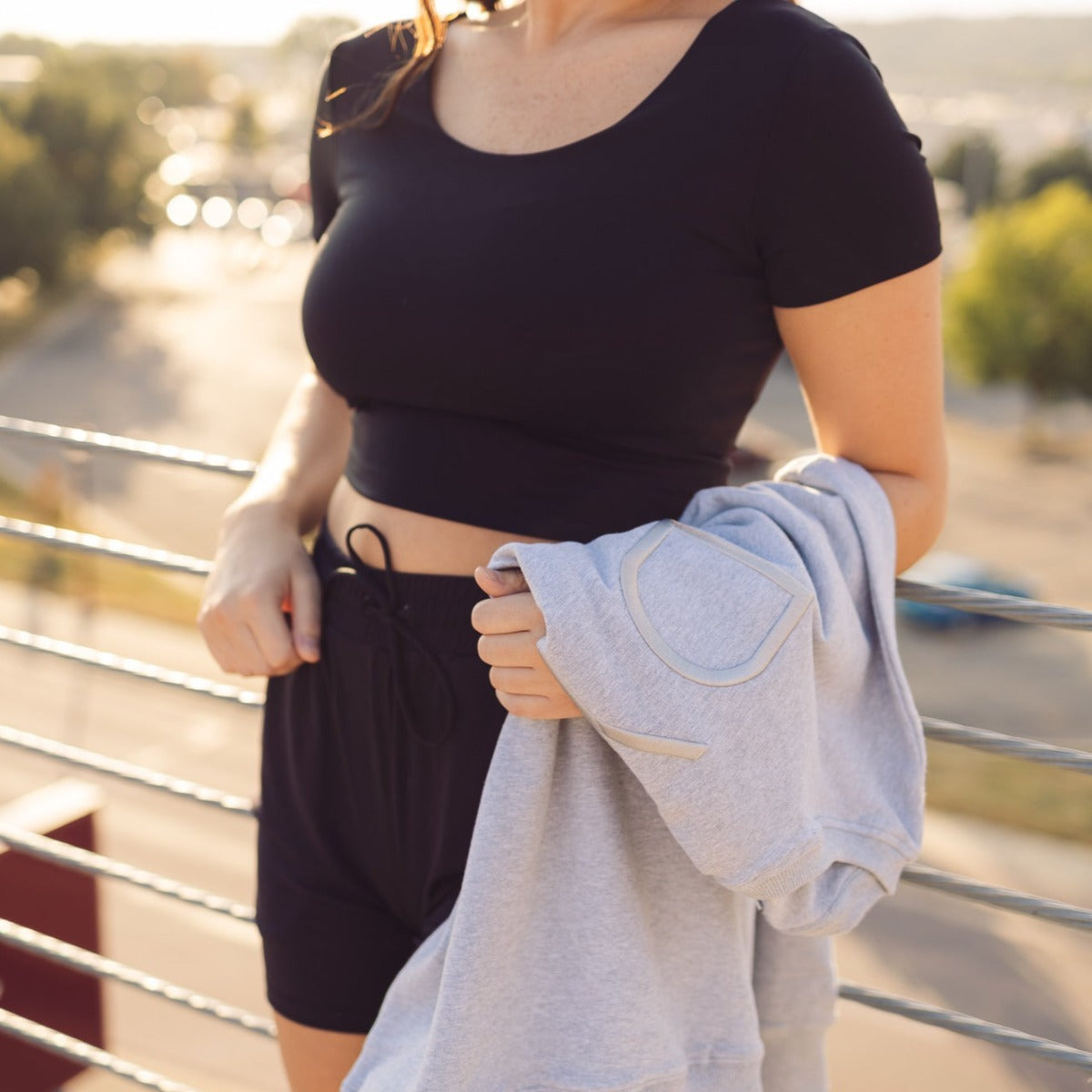 A woman stands outside on a sunny day wearing the DYM Athletics Bliss Two-Way Short Sleeve Top in black with shorts, holding a light gray sweatshirt. The versatile activewear is perfect for warm days, with blurred trees and cars in the background.