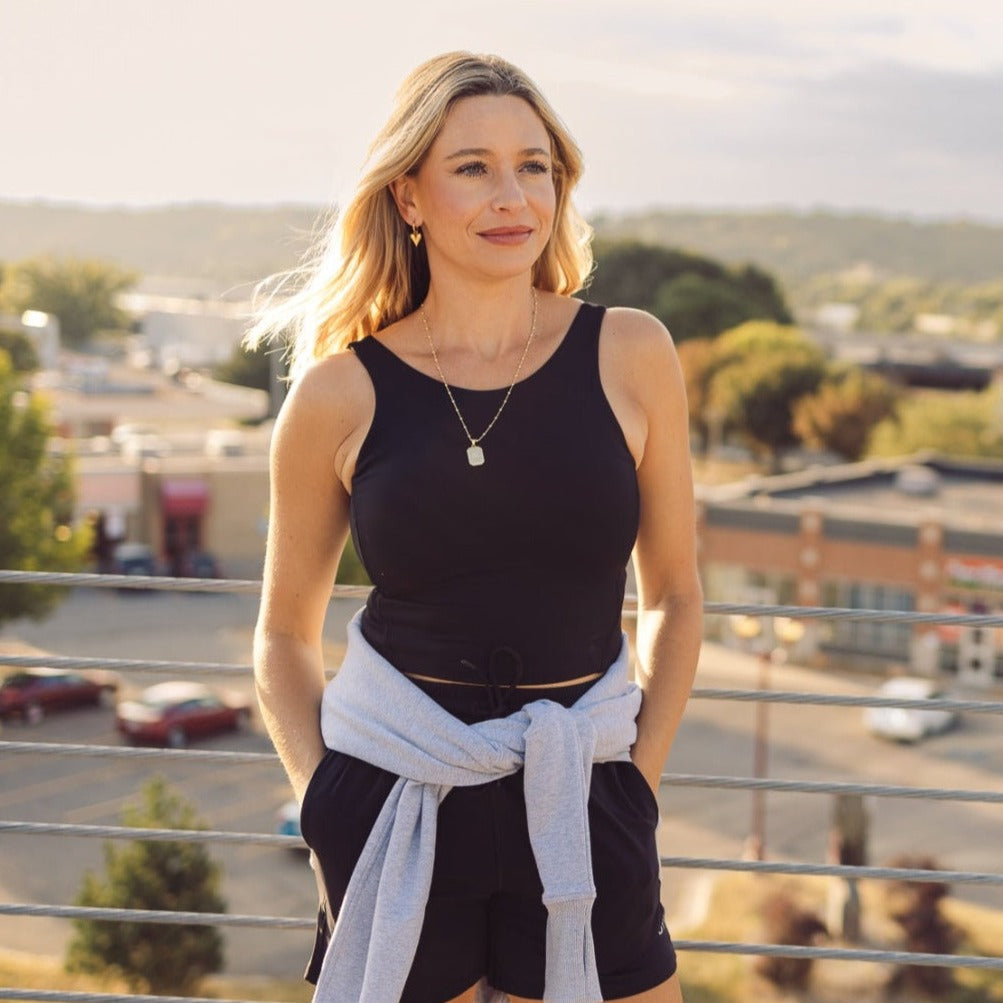 A woman with long blonde hair smiles outdoors, wearing the DYM Athletics Flex High Neck Bra w/ Scoop Back (A-D cups, Medium Support), black shorts, and a light gray sweatshirt around her waist as she leans on a railing before a cityscape.
