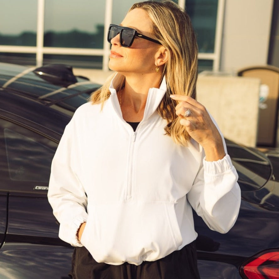 A woman with blonde hair wears black sunglasses and a white Ascend 1/4 Zip Jacket by DYM Athletics. Standing beside a black car, she smiles softly as sunlight highlights her face and hair.
