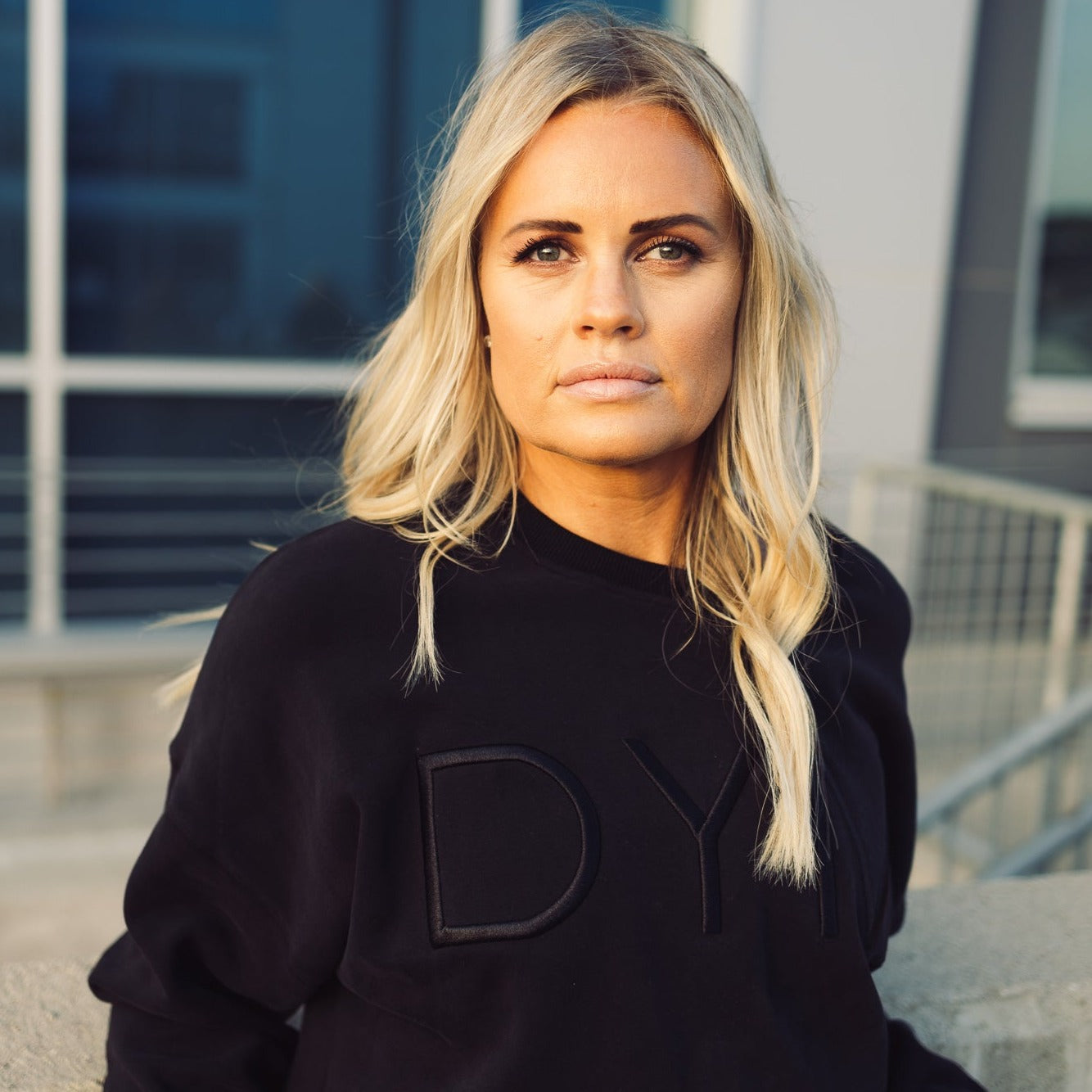 A woman with long blonde hair and a serious expression stands by a metal railing, wearing the Cozy Up Crew Neck Sweatshirt from DYM Athletics. The relaxed fit black unisex top features bold DYM lettering. Glass windows and a modern building blur in the background.