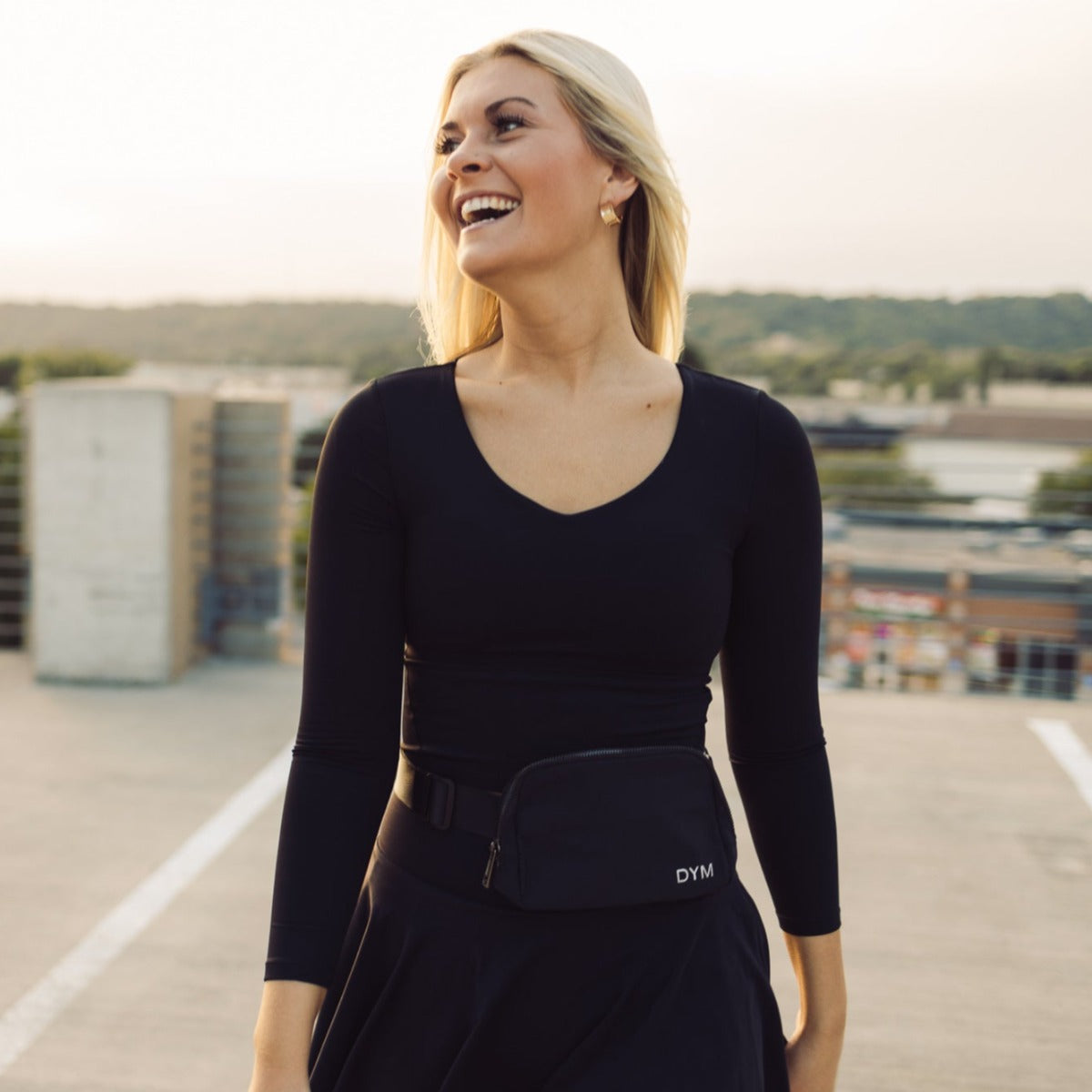 A smiling woman with long blonde hair wears a black DYM Athletics Flex Long Sleeve w/ Built in Bra and a black waist bag labeled “DYM.” She stands outdoors on a rooftop, surrounded by buildings and greenery in soft, natural light.