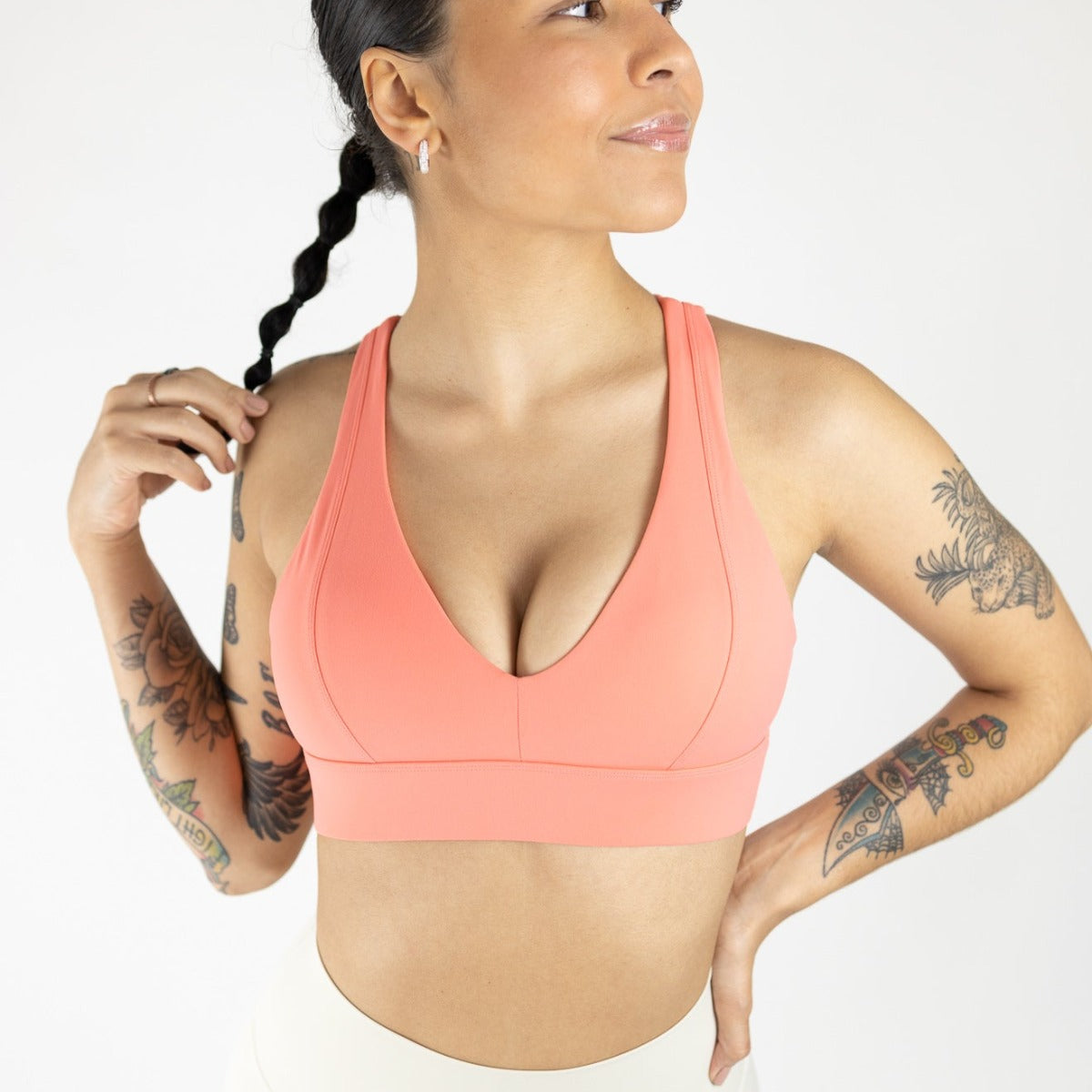 A tattooed woman wears the DYM Athletics Heartbreaker Deep V Sports Bra with Keyhole Back (A-D Cup, High Support) in coral and light leggings. She holds her braided hair, smiling gently against a clean white backdrop.