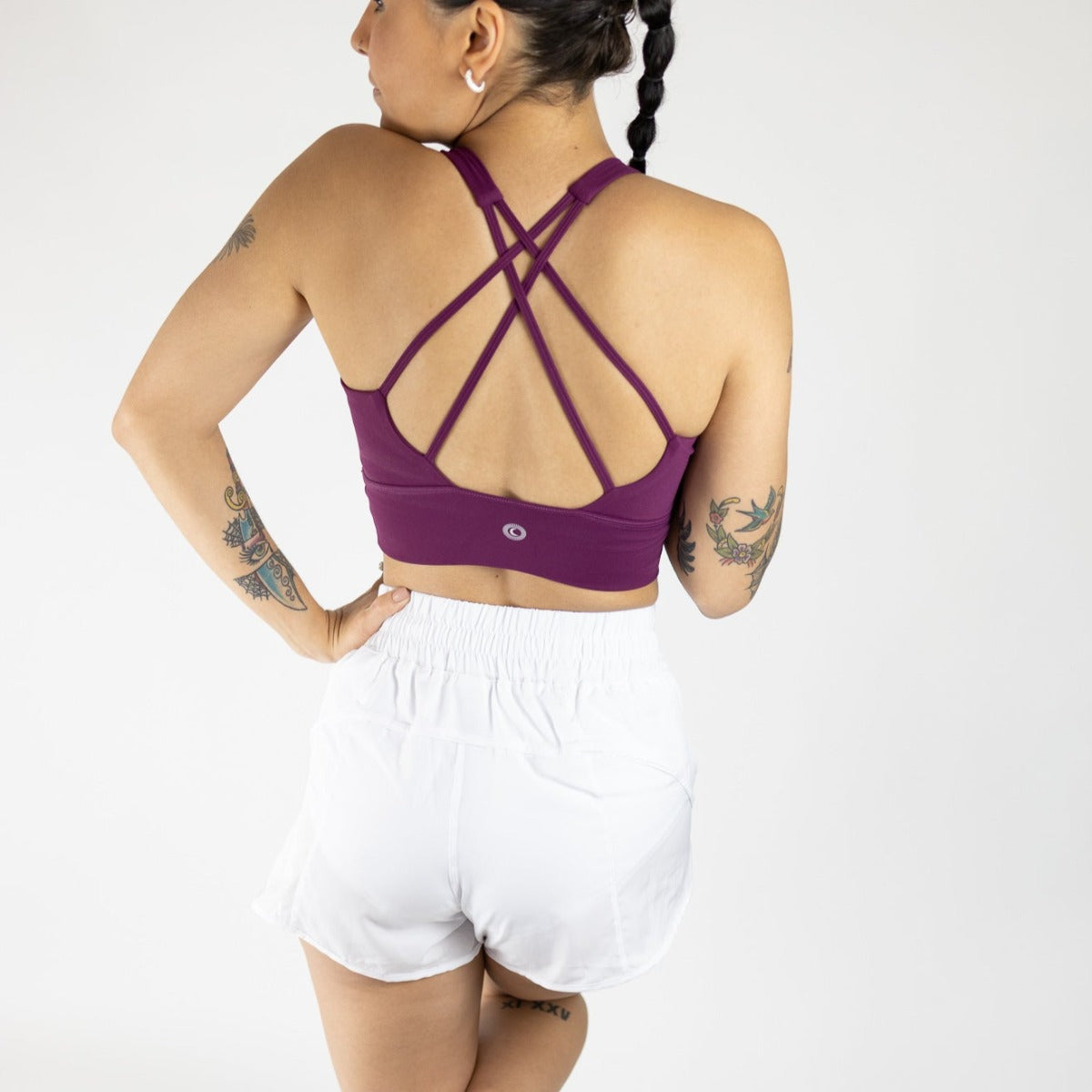 A person with braided hair and medium skin tone stands against a white background, facing away. They wear a purple strappy sports bra and DYM Athletics' Sprint High-Rise Lined Short | 3", made from breathable fabric, with tattooed arms on their hips.