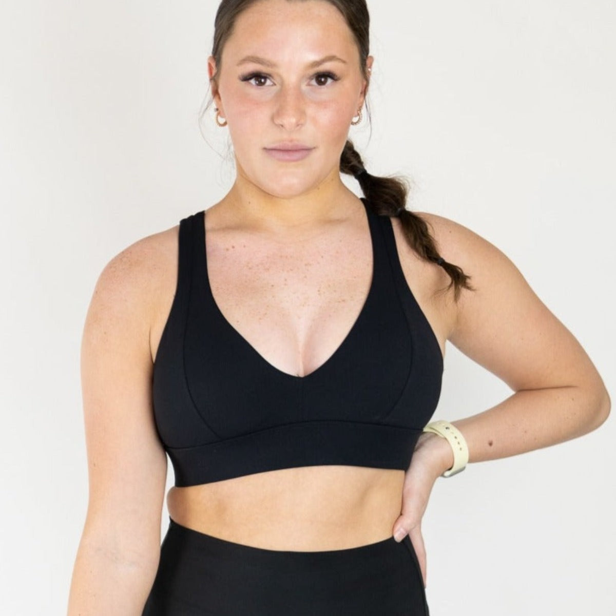 A woman with light skin and brown hair pulled back models the DYM Athletics Heartbreaker Deep V Sports Bra with Keyhole Back (A-D Cup, High Support) in black. She stands confidently against a white background, hand on hip, wearing small hoops and a pale smartwatch.