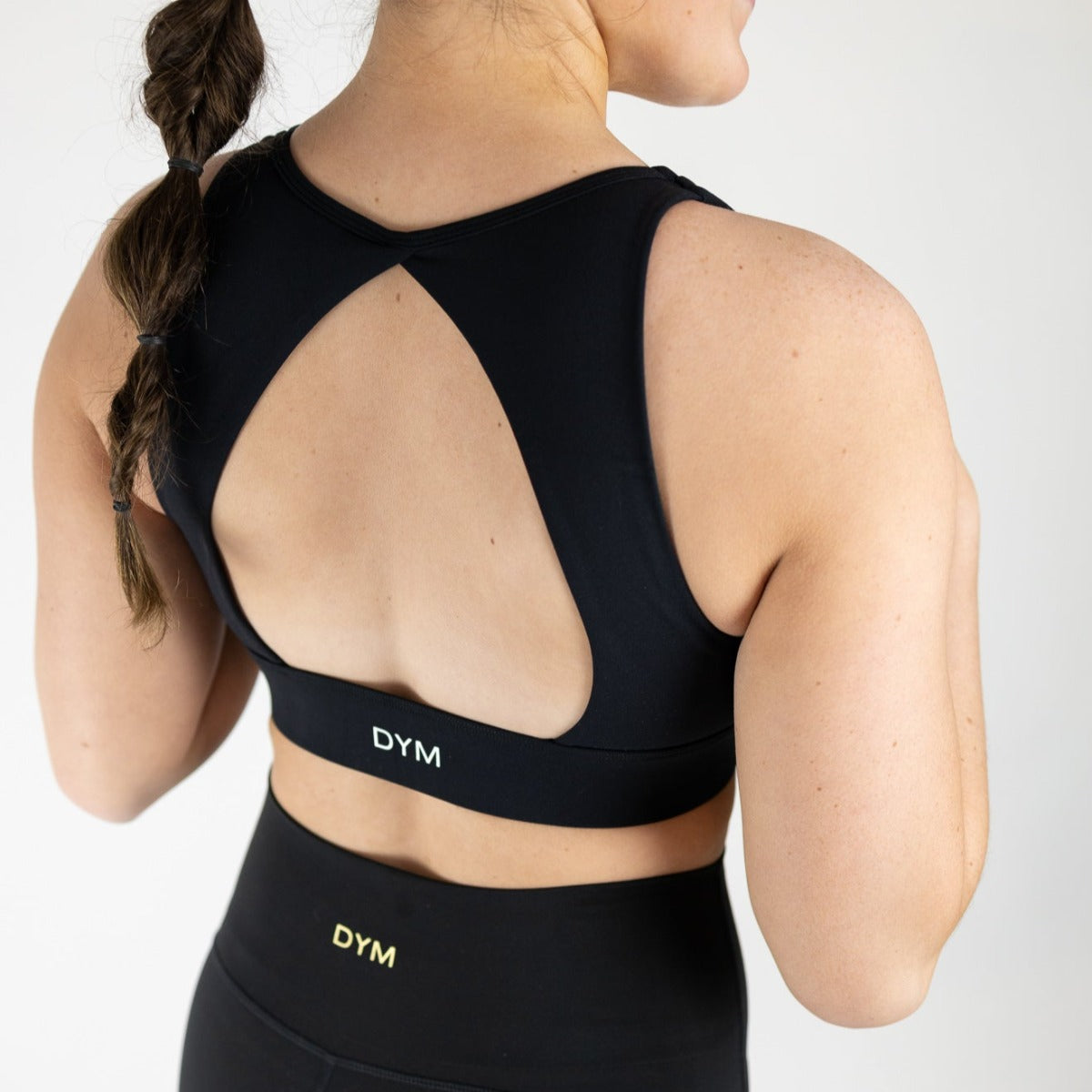 A woman with a braided ponytail wears the Heartbreaker Deep V Sports Bra with Keyhole Back and matching high-waisted leggings by DYM Athletics, both featuring "DYM" in white, as she stands against a plain white background.