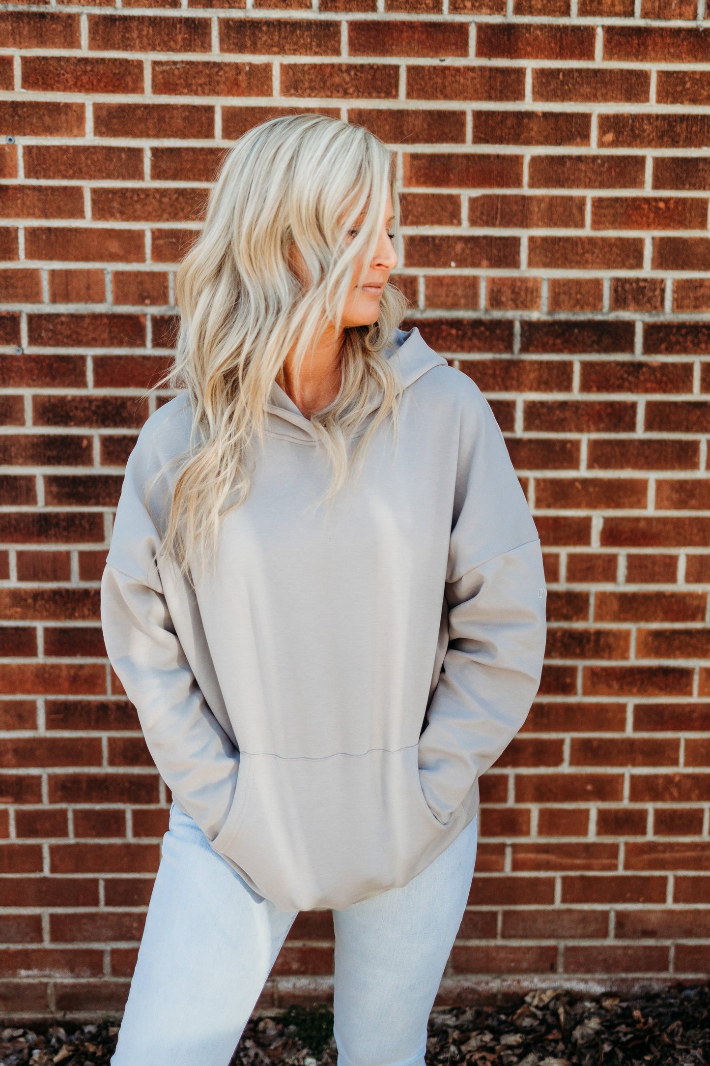A woman with long, wavy blonde hair stands against a brick wall, looking right. She wears the DYM Athletics Cozy Up Boyfriend Hoodie | SALE with light blue jeans, hands in the pocket. The vibe is casual and relaxed.