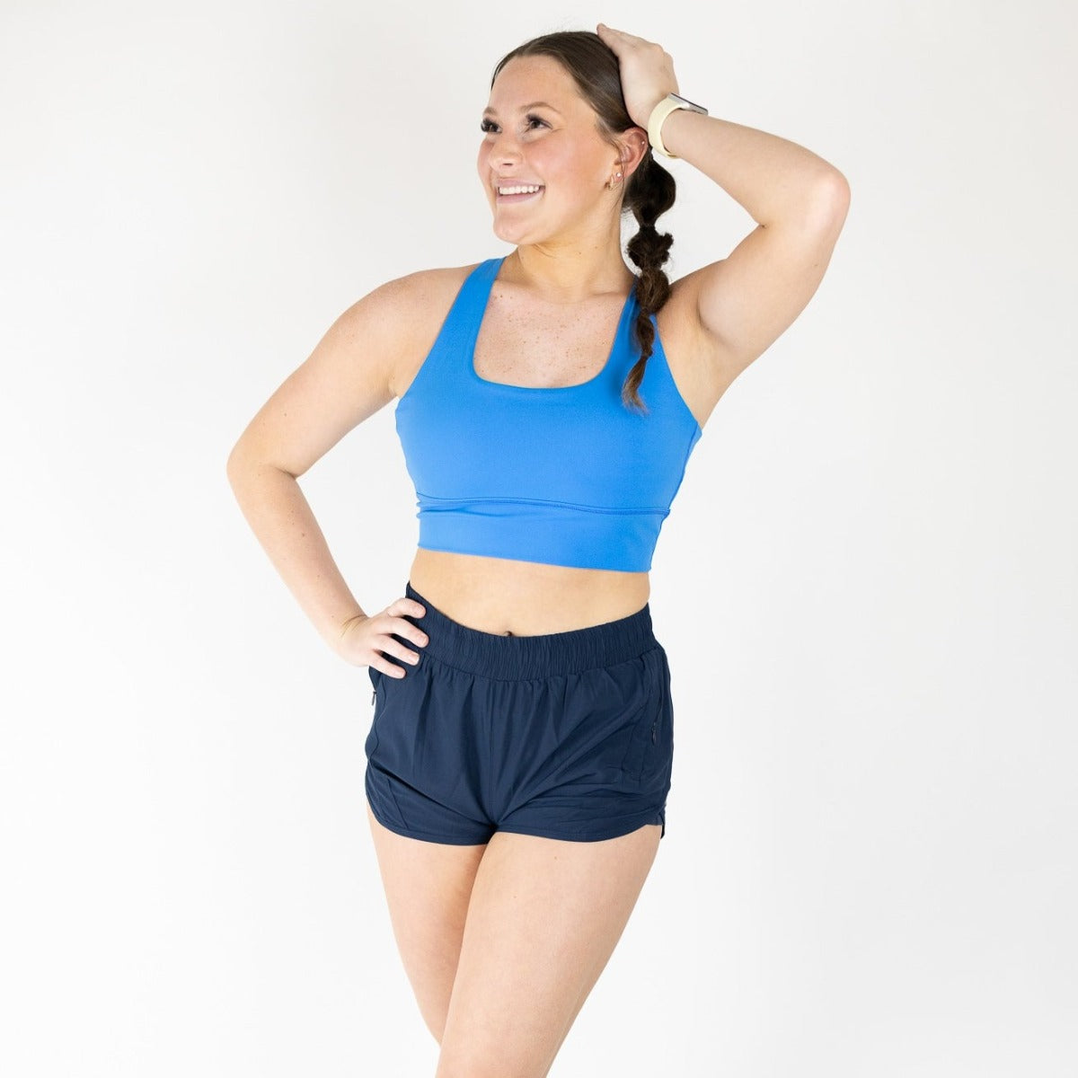 A woman poses confidently against a white background, wearing a blue sports bra and DYM Athletics Speed Mid-Rise Shorts | 2" | SALE in dark blue with zipper pockets. Her hair is in a braid, and she smiles with one hand on her hip.