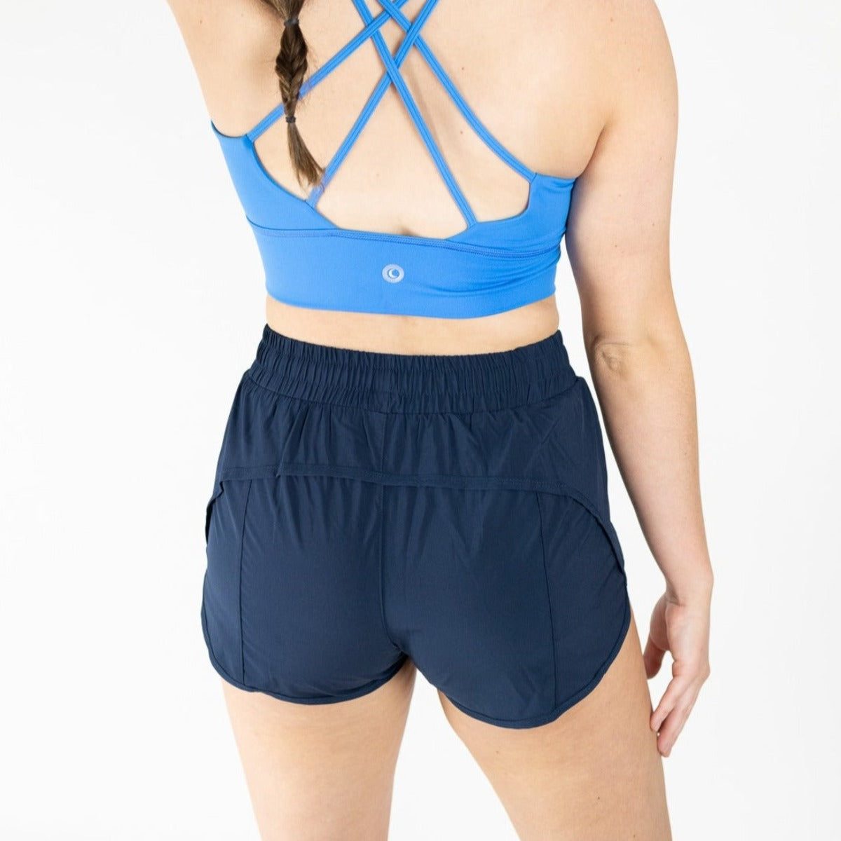 A woman with a single braid stands against a white background, wearing DYM Athletics’ Speed Mid-Rise Shorts | 2" | SALE in dark navy and a blue sports bra with crisscross straps, her arm relaxed at her side.