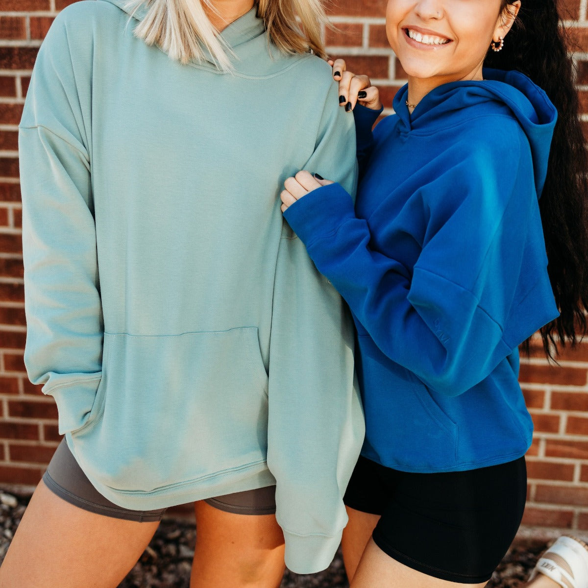 Two women smile side by side in front of a brick wall, wearing DYM Athletics Cozy Up Boyfriend Hoodies—one in light green with gray shorts, the other blue with black shorts. Their faces are partially visible as they hold each other's arms.