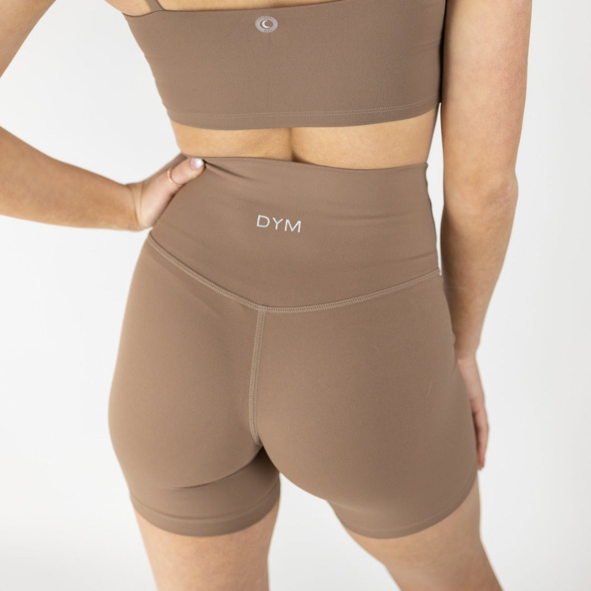 A person stands with their back to the camera, wearing a brown sports bra and DYM Athletics Serene Biker Short | 5" | SALE. One hand is on their hip, and "DYM" is printed in white on the waistband of these super soft shorts. The background is plain white.