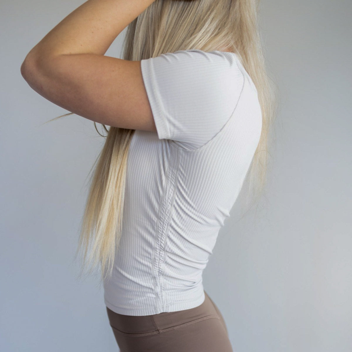 A person with long, straight blonde hair stands in profile, facing left, wearing a fitted cropped Restore Ribbed T-shirt by DYM Athletics and high-waisted brown leggings. Their arms are raised near their head against a plain, light background.