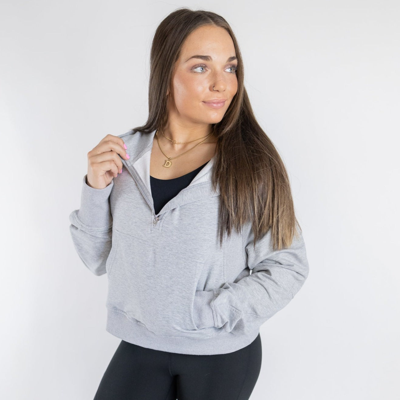 A woman with long brown hair stands against a white background, wearing the DYM Athletics Lightweight 1/4 Zip Hoodie | SALE over a black top and leggings. She holds the zipper, looks to the side, and wears a gold pendant necklace.
