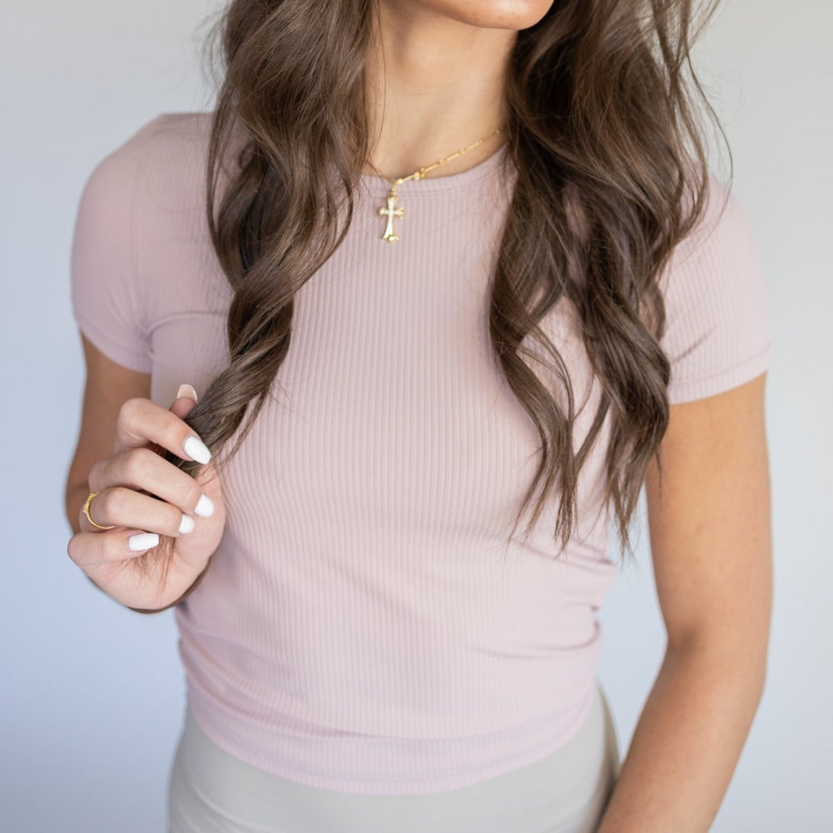 A woman with wavy brown hair wears the DYM Athletics Restore Ribbed T-shirt in light pink, cropped and fitted with short sleeves, paired with beige pants. She accessorizes with a gold ring and cross pendant; her face isn't fully shown.