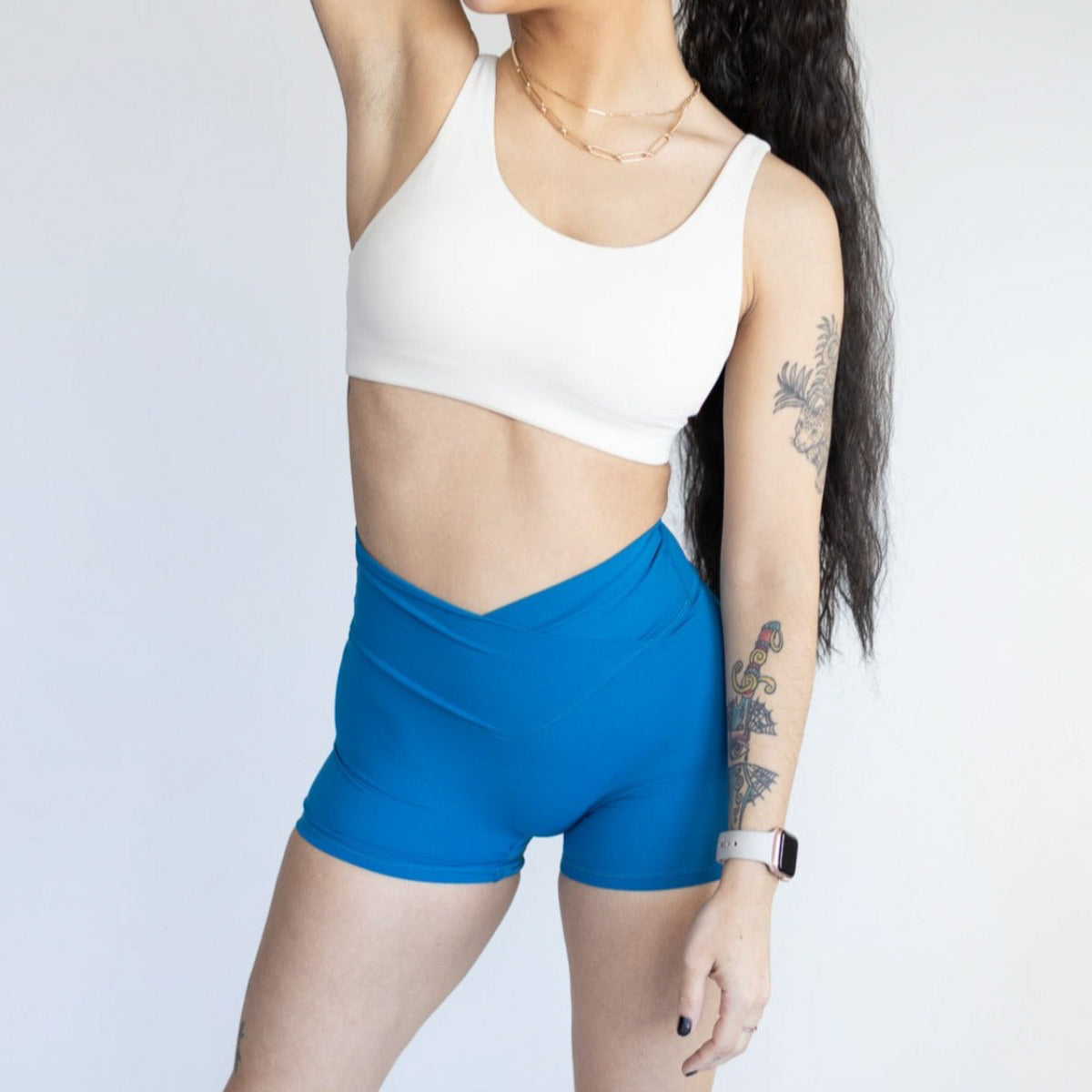 A person with long dark hair wears a white sports bra and DYM Athletics Flex & Flow V-Waist Short | 4" in bright blue. Tattoos are visible on their right arm, a watch on their left, and the background is plain white. Face is mostly out of frame.