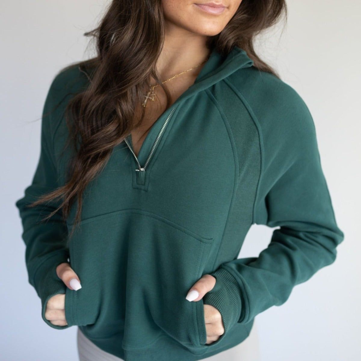 A woman with long brown hair wears the deep green Cozy Up Full Length 1/2 Zip Sweatshirt by DYM Athletics, hands in the front pocket. She accessorizes with a gold necklace; her face is partially visible against a softly lit, plain background.