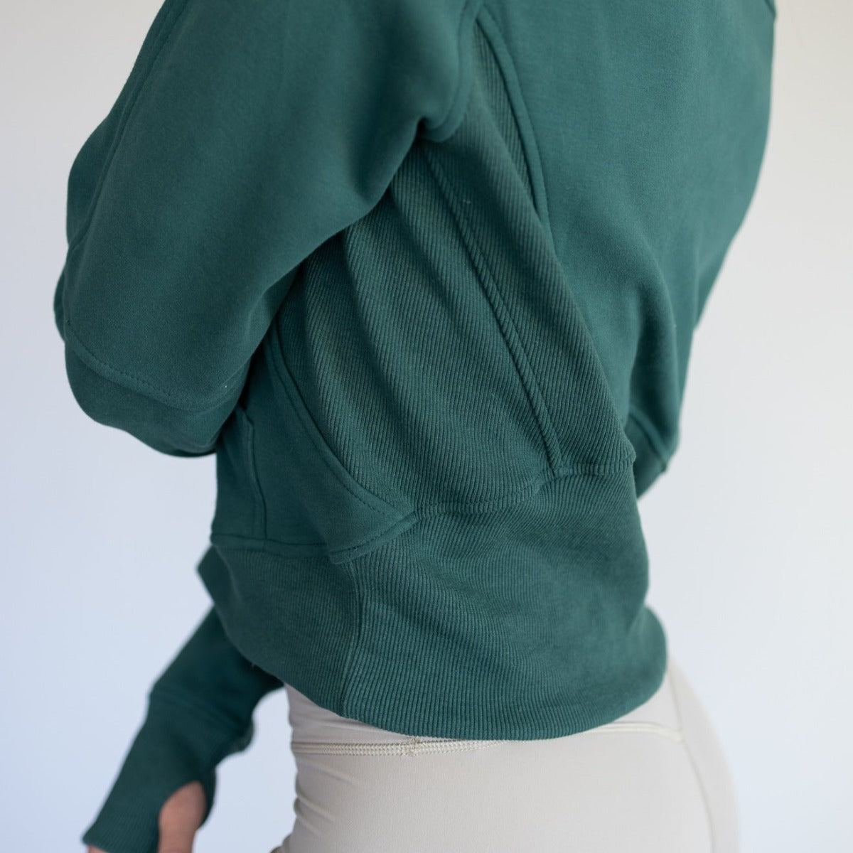 A model wears the DYM Athletics Cozy Up Full Length 1/2 Zip Sweatshirt in dark green with subtle ribbed detailing, paired with light beige fitted pants. The side and back torso view is shown against a softly blurred neutral background.