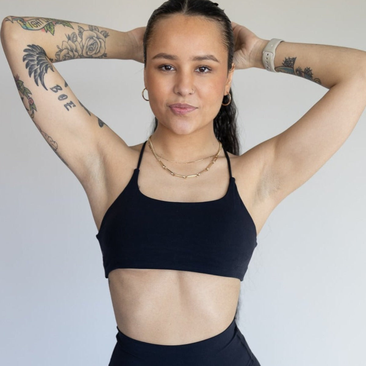 A woman with long dark hair, tattoos, and gold hoop earrings poses with her hands behind her head. She wears the DYM Athletics AIRE Ultra-Soft Open Back Bra for A-C cups (light support), high-waisted leggings, layered necklaces, and a smartwatch.