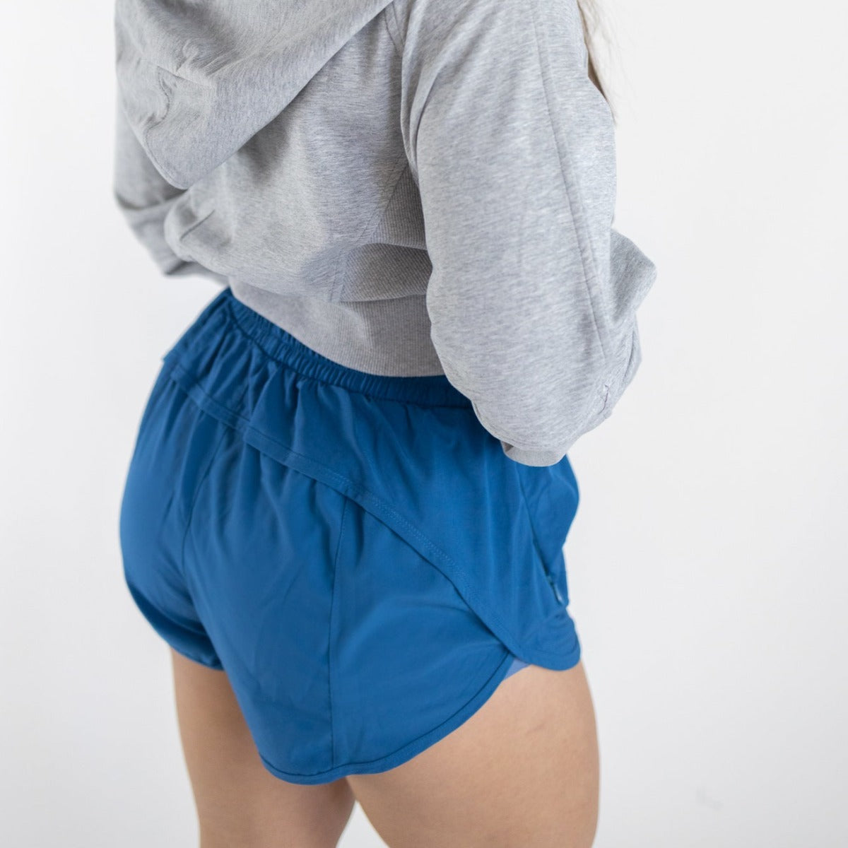 A person faces away from the camera wearing DYM Athletics Speed Mid-Rise Shorts | 2" (SALE) in blue and a light gray hoodie. Only their lower back and upper legs are visible against a plain white background.