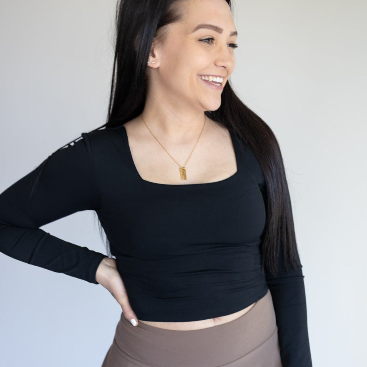 A woman with long dark hair and fair skin smiles while looking left, wearing the DYM Athletics Bliss Square Neck Long Sleeve Top | SALE with brown high-waisted pants and a gold necklace, one hand on her hip against a plain light background.