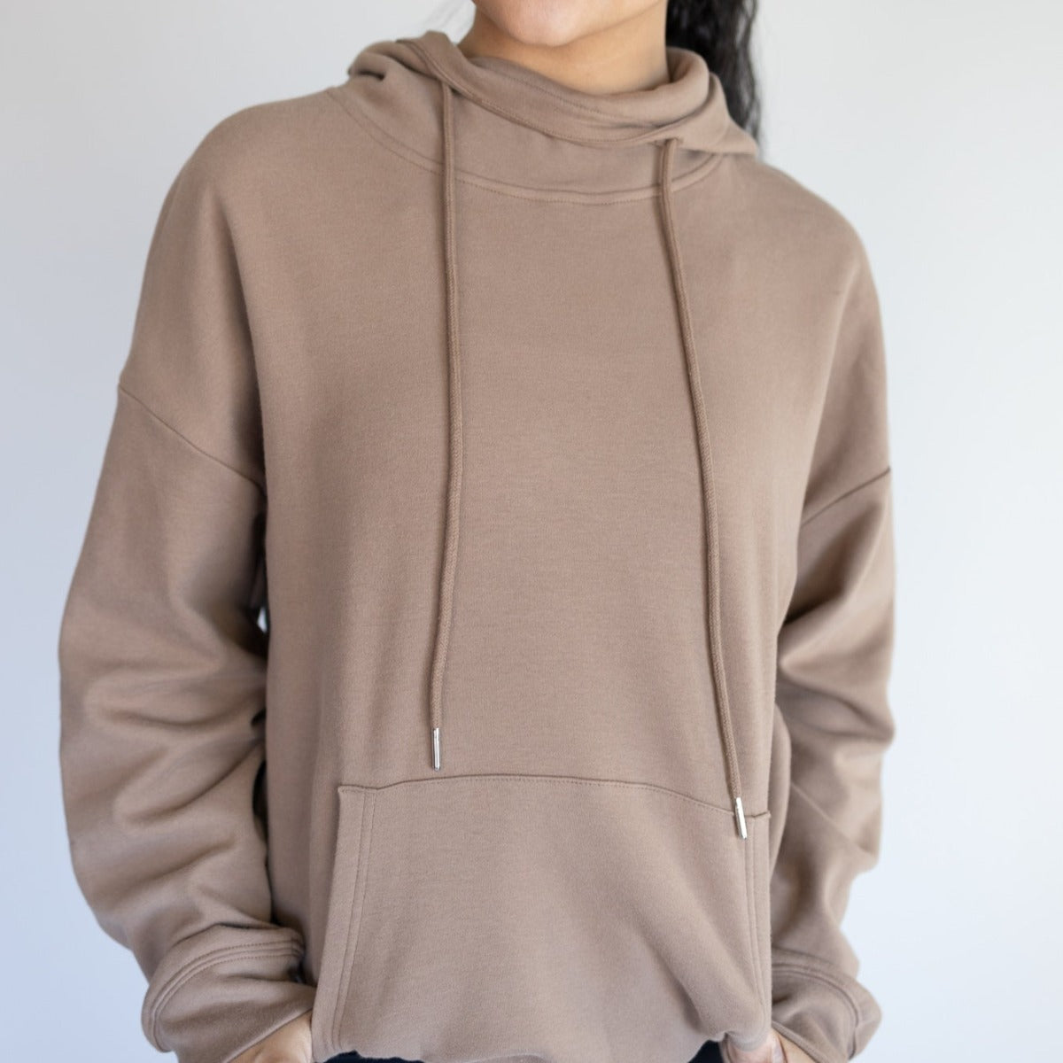 A person wears the DYM Athletics Cozy Up Cowl Neck Hoodie, an oversized light brown sweatshirt with a front pocket, drawstrings, long sleeves, and an ultra soft feel. Their hands are partially in the pocket against a plain light background.