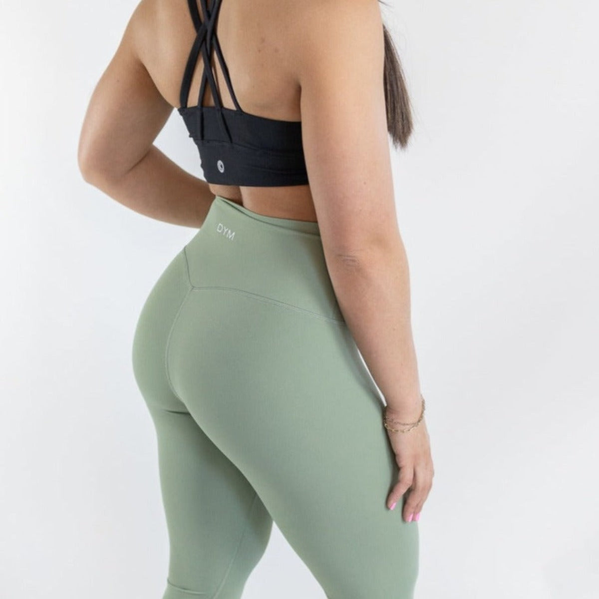 A woman, seen from behind, wears DYM Athletics' Ultra High Waist Flex & Flow Legging in sage green with a black sports bra. Her long brown hair drapes over her shoulder against a plain white background.