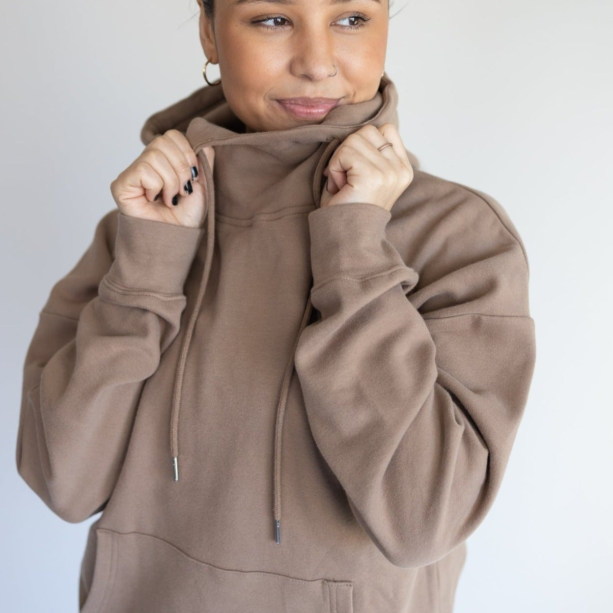 A person smiles softly, holding up the collar of a DYM Athletics Cozy Up Cowl Neck Hoodie in brown, partially covering their mouth. They sport black nail polish and a small nose ring against a plain light background.