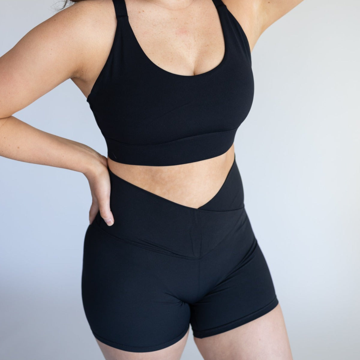 A woman models DYM Athletics' Flex & Flow V-Waist Short | 4" (SALE) in black with a matching sports bra. She stands against a light background, left hand on hip, right arm bent behind her head; her face is not shown.