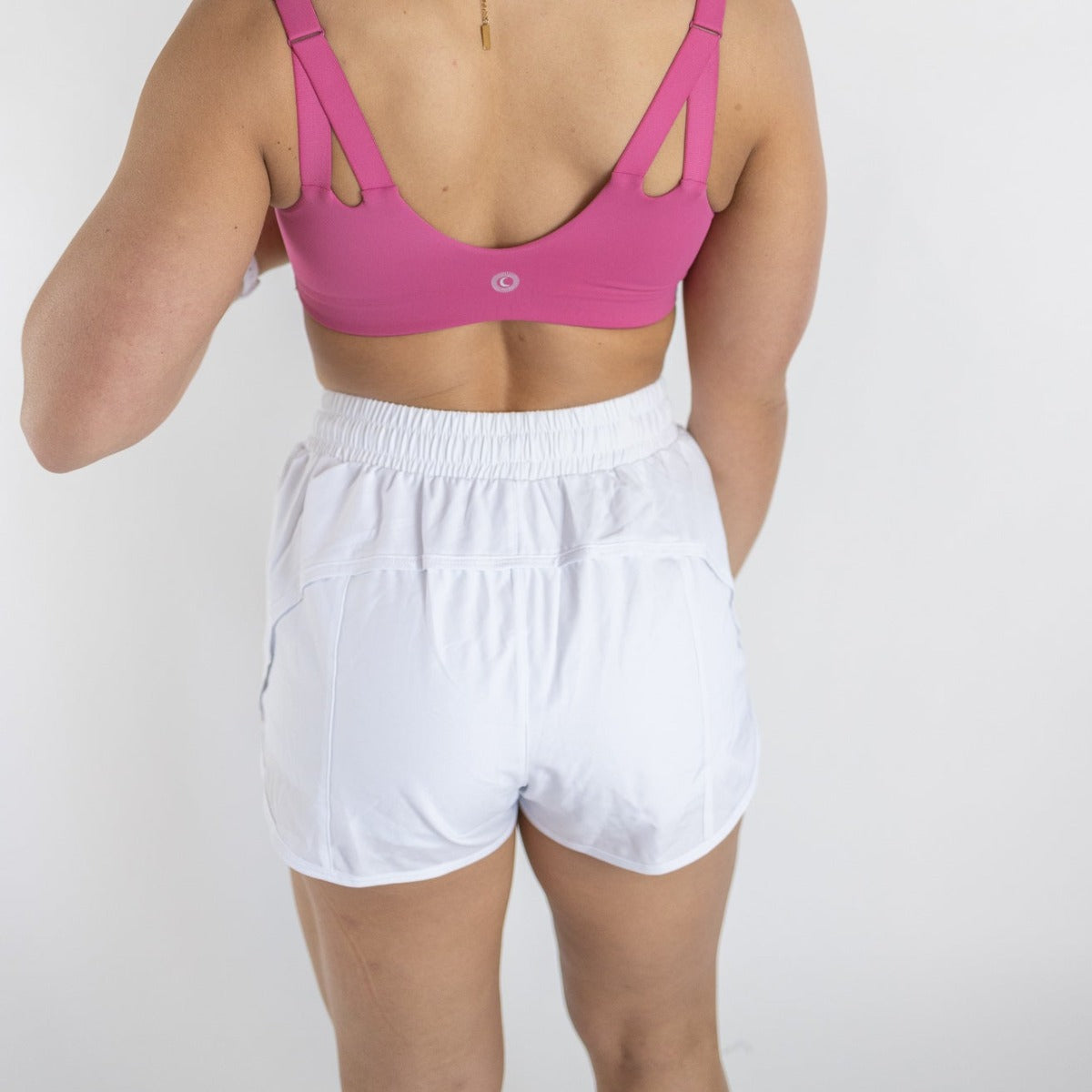 A person stands facing away, wearing a pink sports bra and DYM Athletics Speed Mid-Rise Shorts | 2" | SALE with zipper pockets. Their right arm is bent, and the plain white background highlights the workout clothing.