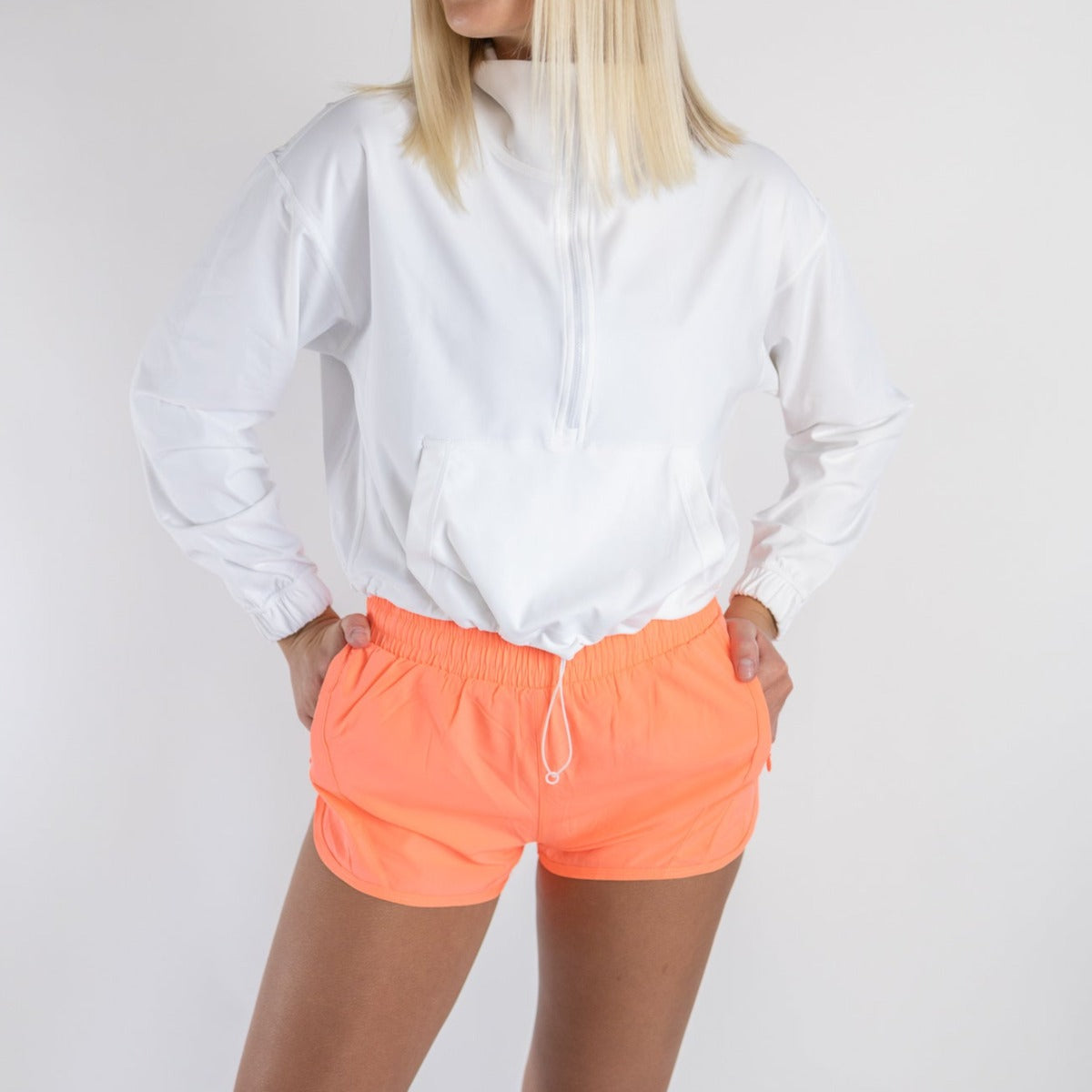 A person with straight blonde hair stands against a white background, wearing a white long-sleeve pullover and DYM Athletics Speed Mid-Rise Shorts | 2" | SALE in bright neon orange, hands on hips and face partially out of frame.