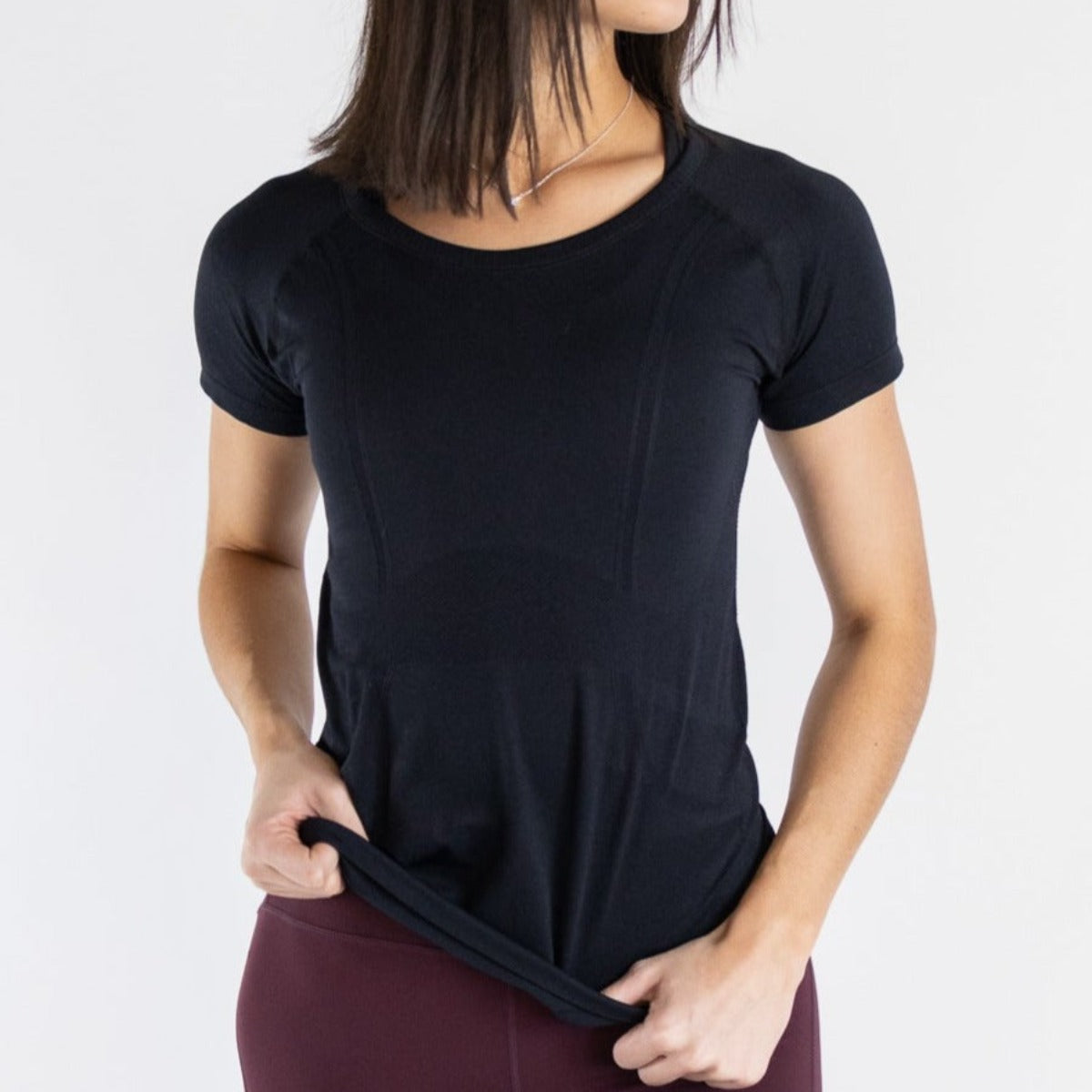 A woman with straight, shoulder-length dark hair wears the DYM Athletics Trainer Lightweight Short Sleeve Shirt in black and maroon leggings. She holds the shirt's hem with both hands and stands at an angle against a plain white background.