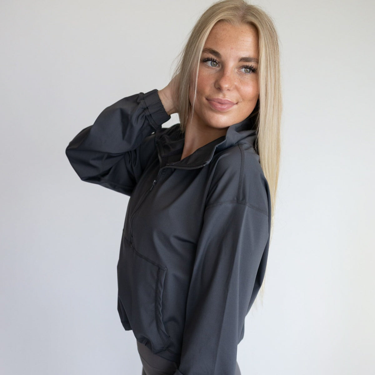 A young woman with long blonde hair and fair skin wears the DYM Athletics Ascend 1/4 Zip Jacket in dark gray. She smiles gently, facing slightly to the side with one hand in her hair, against a plain white background.