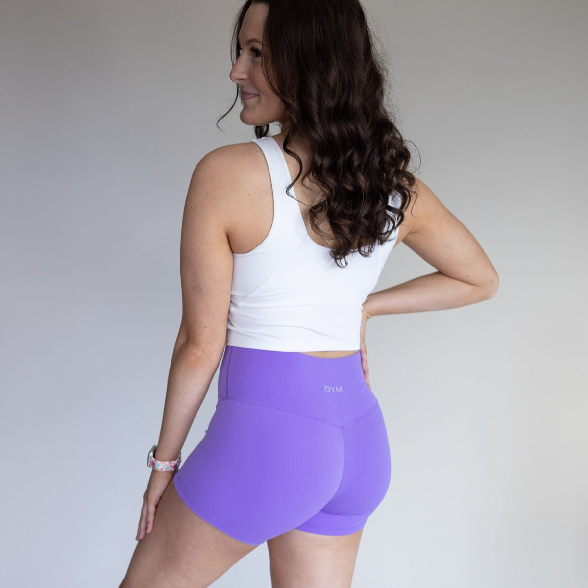 A woman with long, wavy brown hair smiles sideways with one hand on her hip, wearing the DYM Athletics Flex High Neck Bra w/ Scoop Back (A-D cups, Medium Support) and high-waisted purple athletic shorts against a plain background.