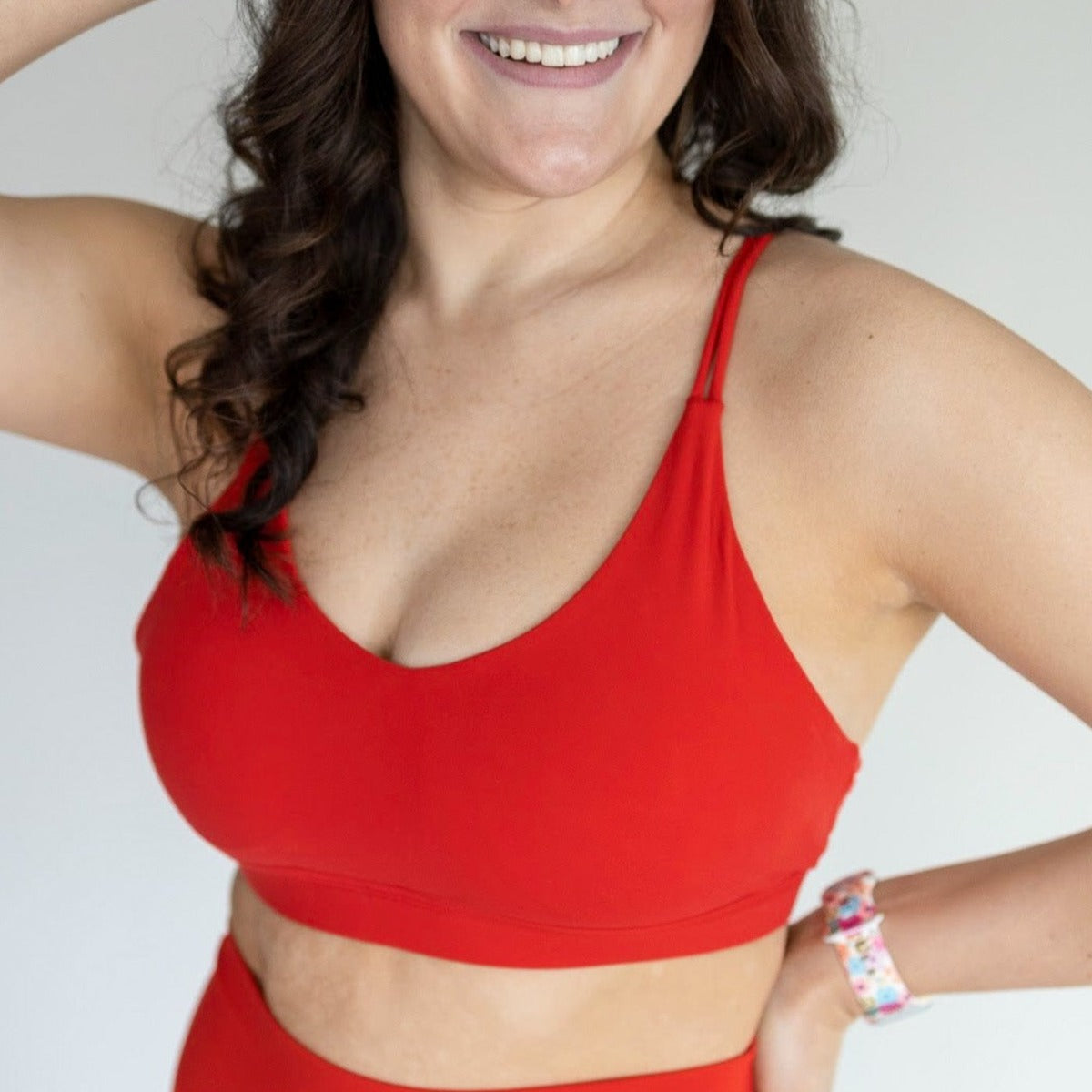 A woman with long brown hair smiles, wearing the DYM Athletics AIRE Ultra-Soft Cross Back Sports Bra (A-D Cup, Medium Support, SALE) in bright red with matching high-waisted bottoms. She poses with a beaded bracelet against a light background.