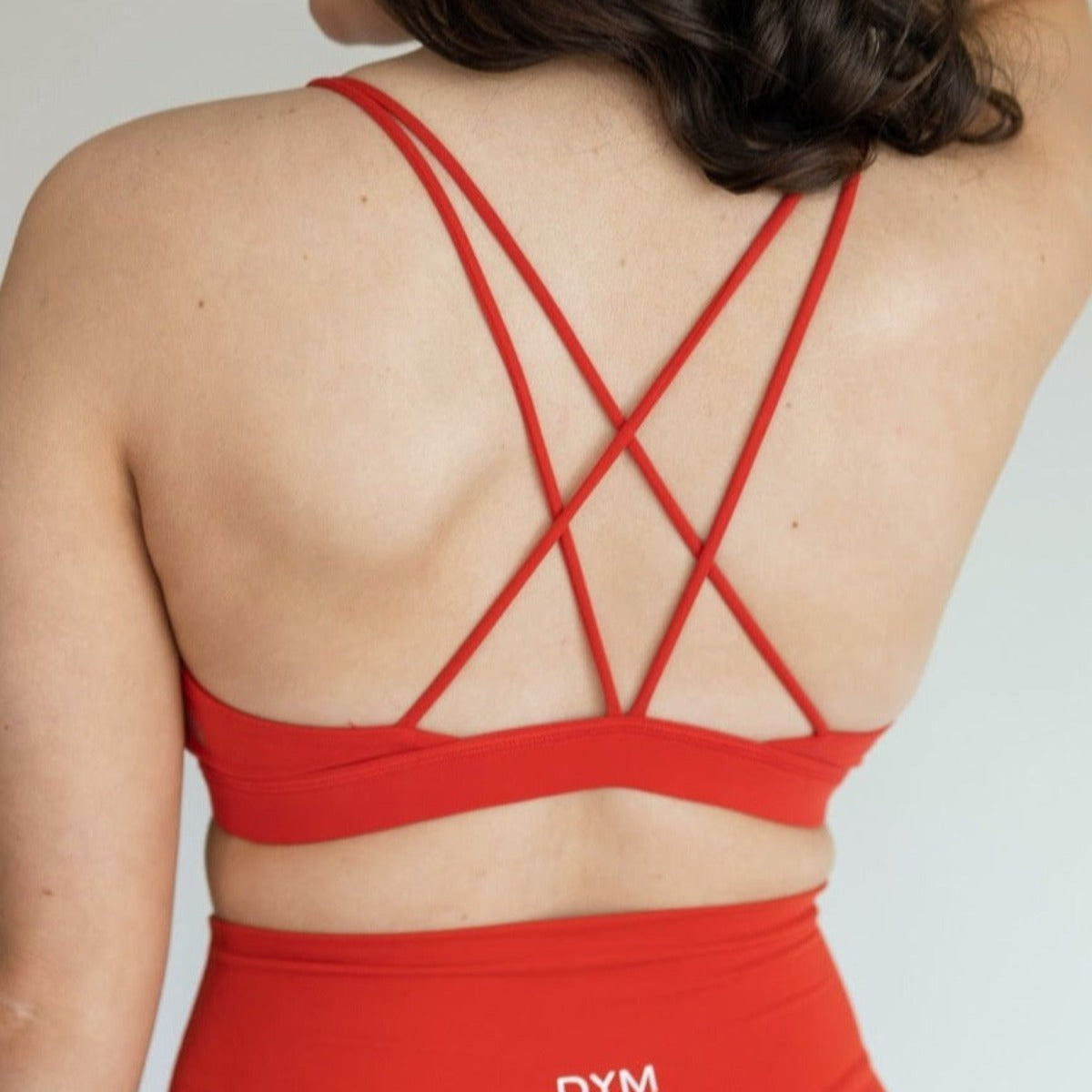 A woman with wavy brown hair is shown from the back, wearing the DYM Athletics AIRE Ultra-Soft Cross Back Sports Bra (A-D Cup, Medium Support) in bright red, paired with matching high-waisted leggings against a plain light background.