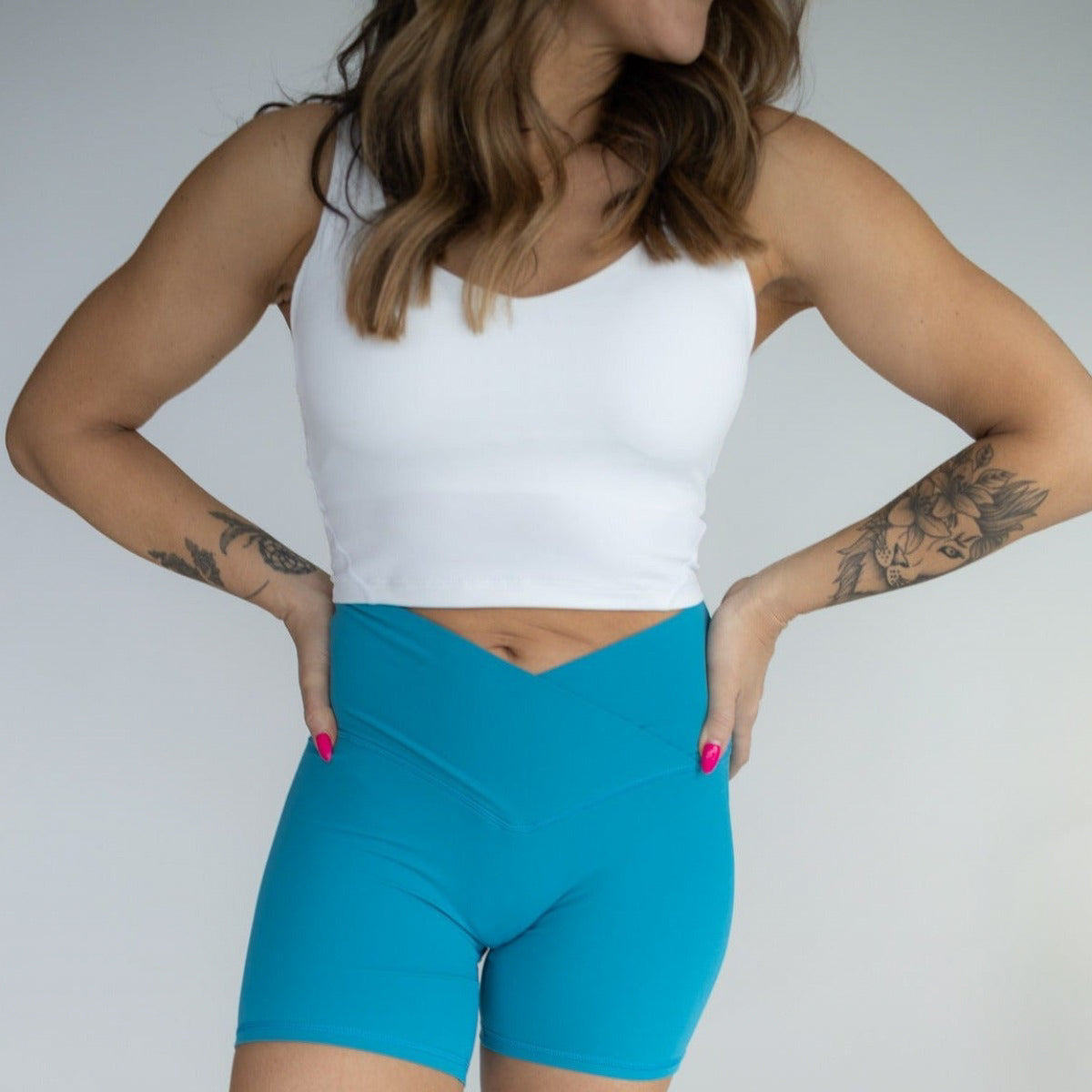 A woman with loose brown wavy hair, pink nails, and forearm tattoos stands hands-on-hips, wearing a white crop top and DYM Athletics' Flex & Flow V-Waist Short | 4" (bright blue). Her face is mostly out of frame.
