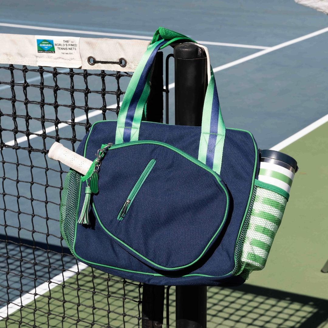 The Fresh Pickle Lola Pickleball Tote Bag in navy with green and white striped handles hangs on a tennis net post. A racket handle peeks out from the side, while a striped water bottle fills the mesh pocket courtside.