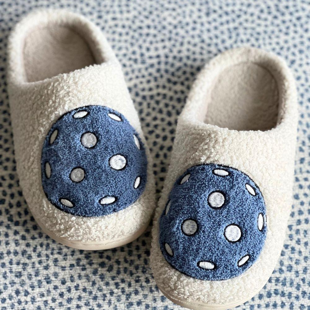 Pickleball Slippers by Fresh Pickle: cozy, cream-colored slippers with a soft, fuzzy texture. Each has a blue patch with white polka dots and stitched details, displayed on patterned blue-and-white fabric.