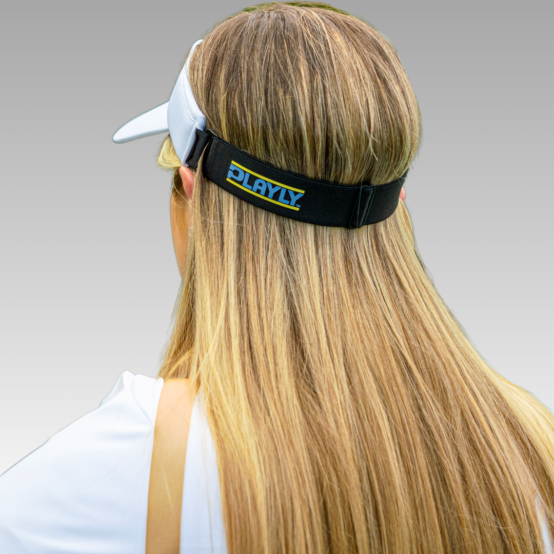 A person with long, straight blonde hair is seen from behind wearing the Playly Store Playly Pickleball Visor, featuring a white UV-protective design and an adjustable black strap with a blue and yellow PLAYLY logo, set against a smooth gray gradient background.