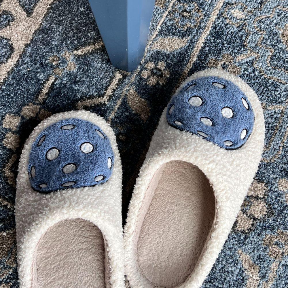 A pair of Fresh Pickle Pickleball Slippers in fuzzy beige, featuring blue and white polka-dotted mushroom cap designs on the toes, sits on a blue patterned rug beside a blue table or chair leg.