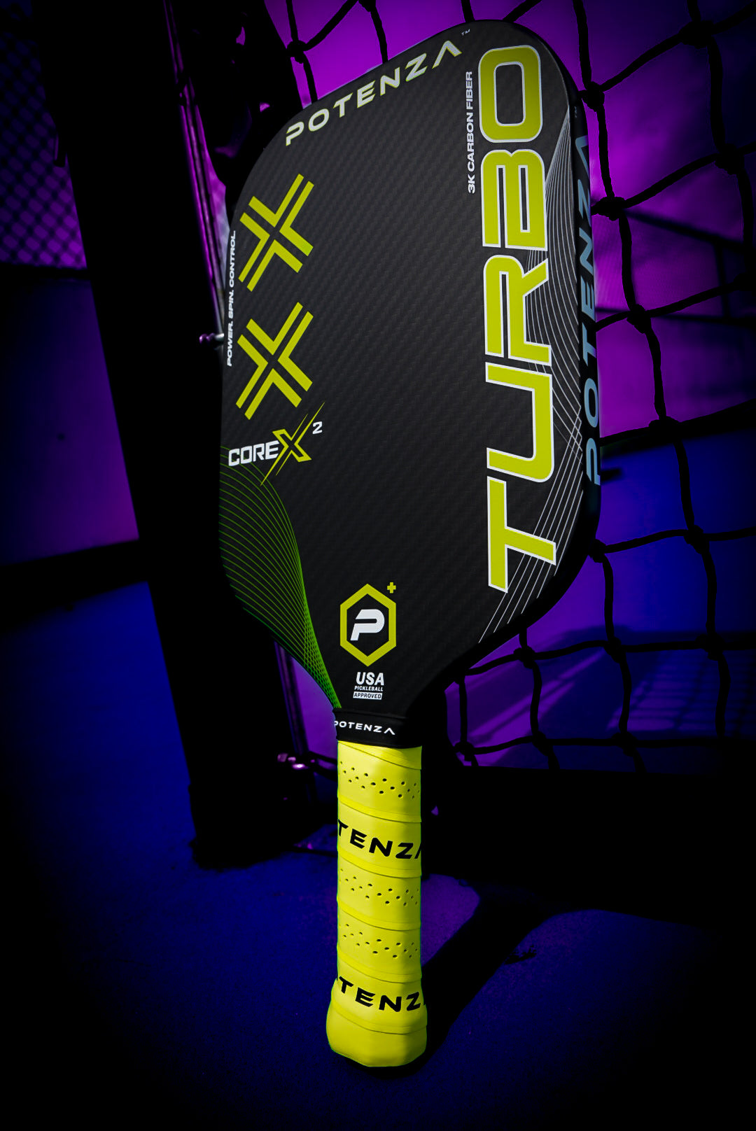 A black Potenza Pickleball TURBO+ COREx2 carbon fiber paddle with neon yellow accents leans against a net on a blue court under purple lighting. The bright yellow grip is labeled POTENZA and the USA Pickleball approved logo is visible.