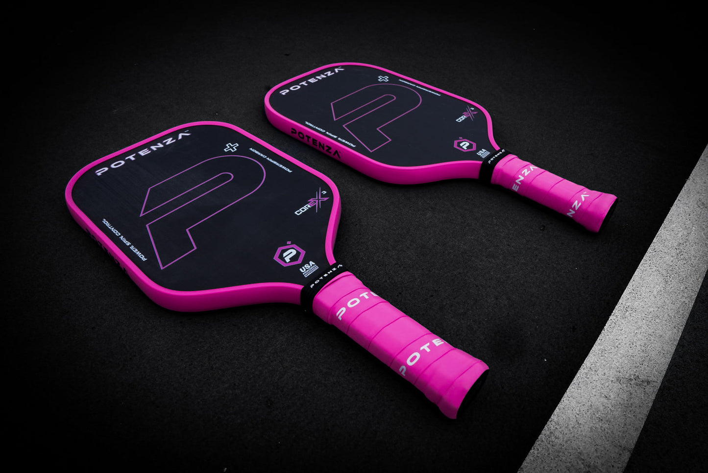 Two Potenza Pickleball P+ PowerSpin Carbon COREx3 (Neon Pink) paddles with black carbon fiber, neon pink edges and handles, plus bold white and purple graphics, are placed side by side on a dark court surface.