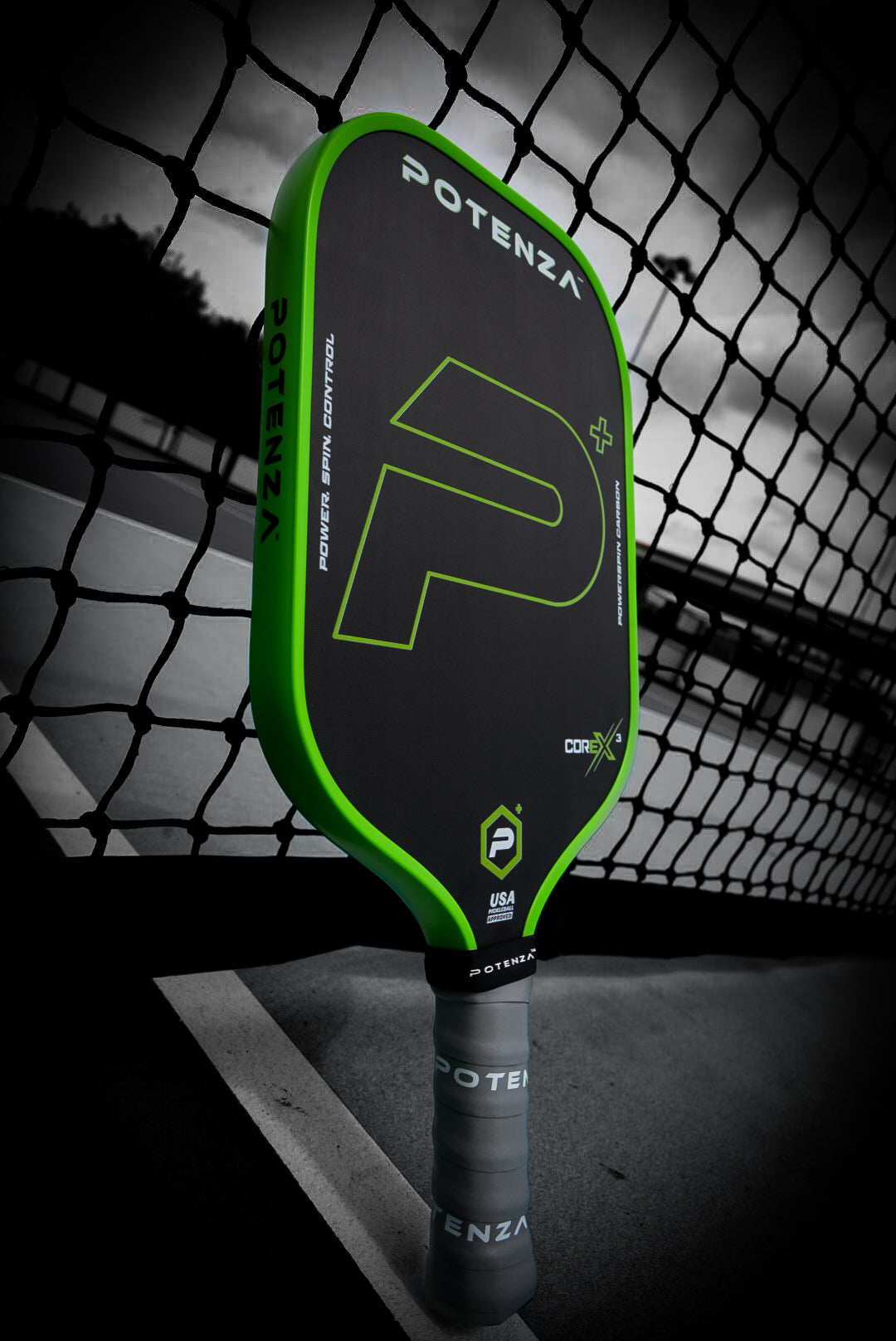 A Potenza Pickleball P+ PowerSpin Carbon COREx3 (Neon Green, Elongated Face) paddle stands upright by a tennis net, showing its large P logo, neon green edges, and branded face and grip against a blurred court backdrop.