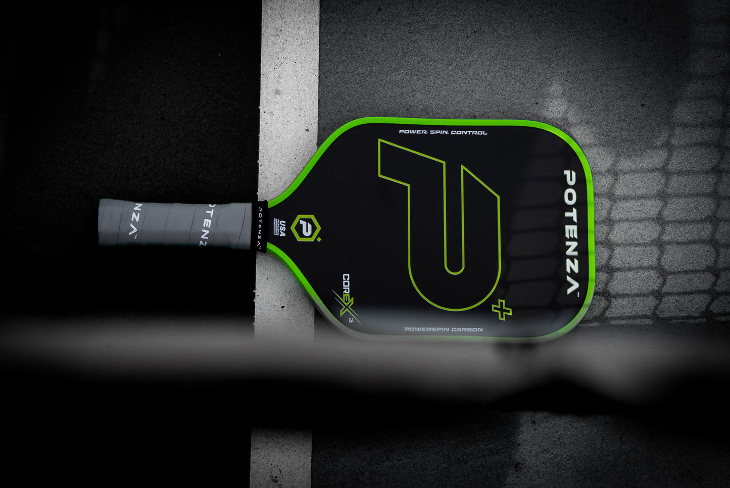 A Potenza Pickleball P+ PowerSpin Carbon COREx3 (Neon Green) paddle with a black and neon green design lies on a dark court, partly over a white line. Its handle features gray grip tape, while geometric shadows cover the lower part of the scene.
