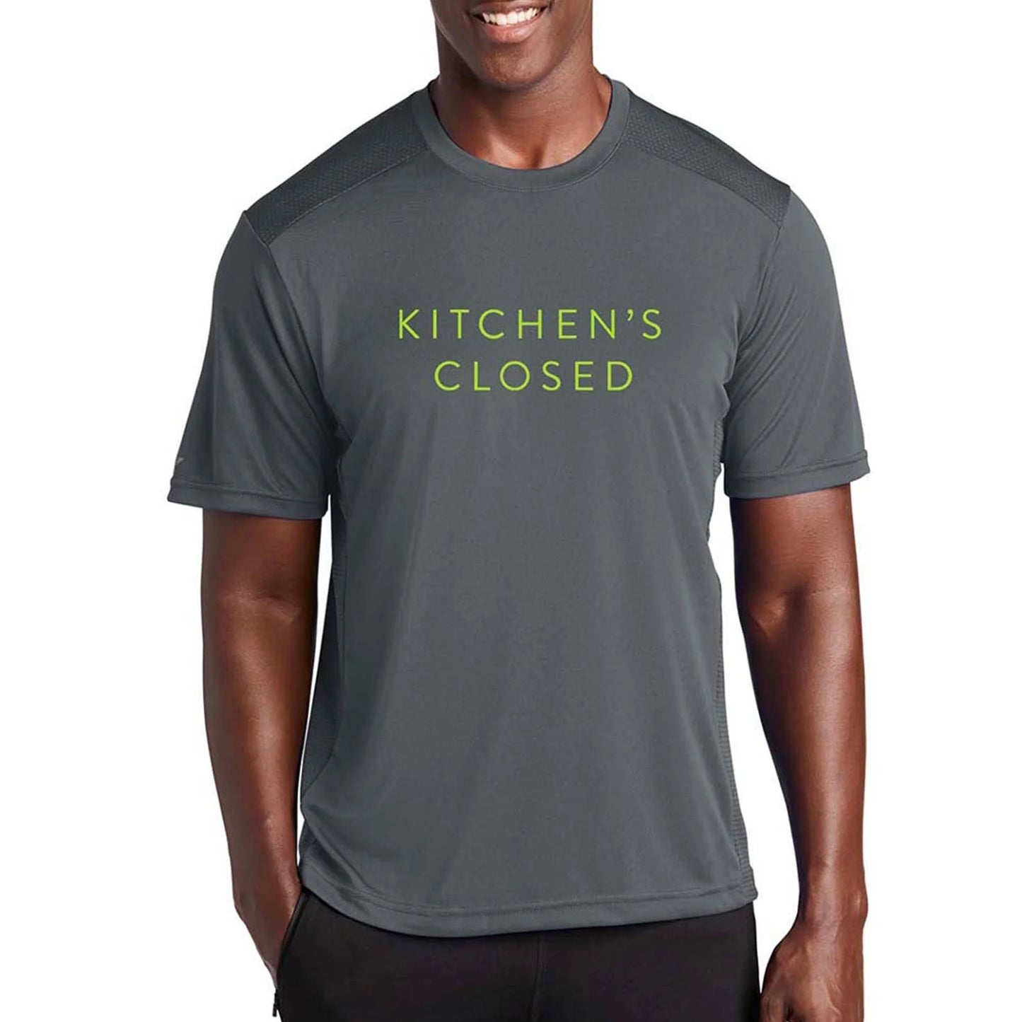 A smiling man wears the Swinton Pickleball Men's Kitchen's Closed Performance Shirt Gray, featuring moisture-wicking fabric and “KITCHEN’S CLOSED” in light green letters—adding a fun pickleball touch. Plain white background highlights its sporty style.