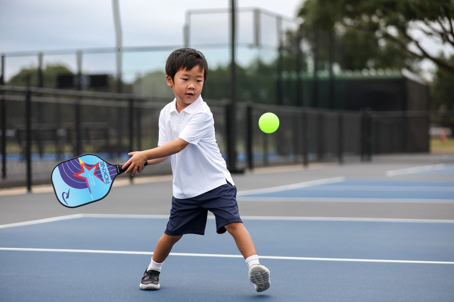 A young boy in a white polo and navy shorts plays pickleball on an outdoor court, swinging the ROKNE CURVE CLASSIC ROK-STAR (KIDS PADDLE) to hit a green ball against a backdrop of chain-link fence, blue courts, and trees in a sports complex.