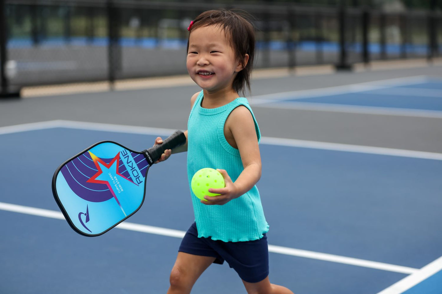 A smiling young girl with short dark hair and a pink hairclip holds the ROKNE CURVE CLASSIC ROK-STAR kids paddle and a neon yellow pickleball on an outdoor court, wearing a turquoise sleeveless top and navy shorts, ready for a fun game.