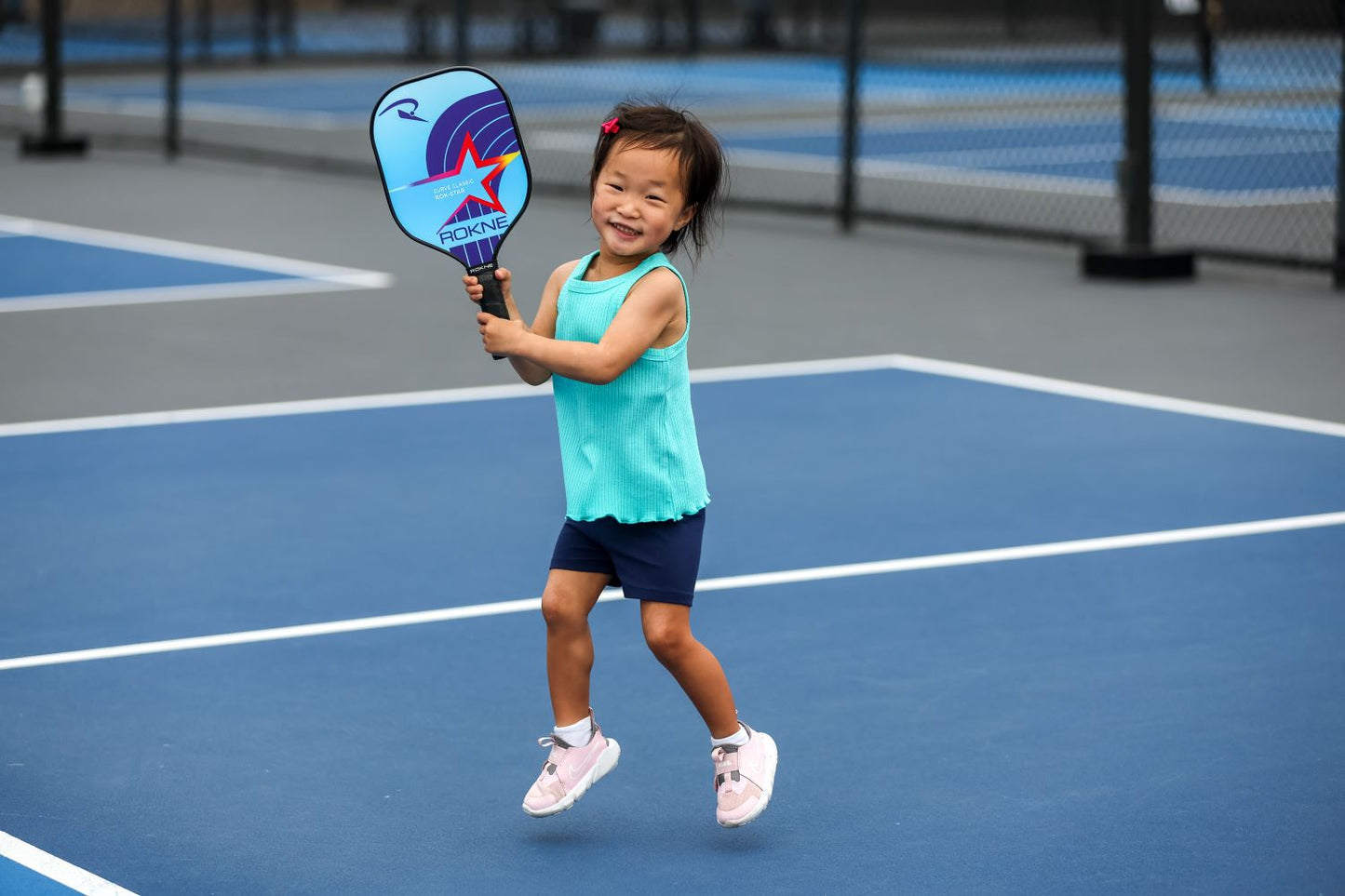 A young girl with short dark hair jumps joyfully on an outdoor pickleball court, smiling brightly as she holds a ROKNE CURVE CLASSIC ROK-STAR (KIDS PADDLE), her pink sneakers and bow standing out against the blue courts.