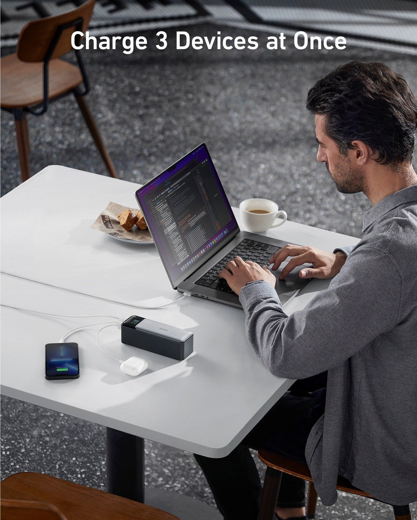 A man works on a laptop at a white café table. An Anker 737 Power Bank (PowerCore 24K) charges his smartphone and earbuds with 140W Power Delivery. Text reads “Charge 3 Devices at Once.” Pastries and a mug are next to the laptop.