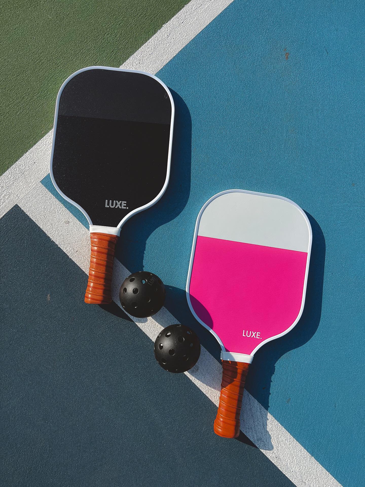 Two LUXE Pickleball Dipped - Leisure paddles with brown handles—one black and white, the other gray and pink—lie on a blue and green court with two black perforated pickleballs between them.