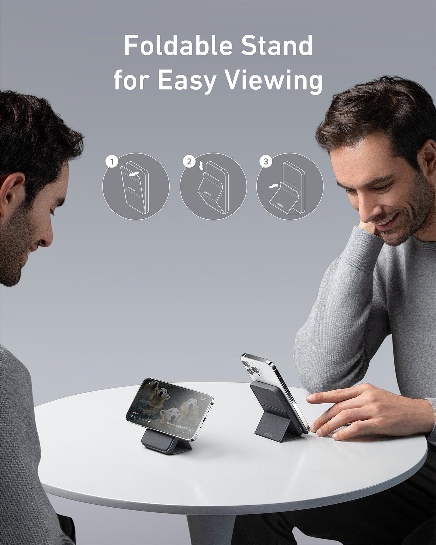 Two men sit at a round white table, each using an Anker 622 Magnetic Battery (MagGo) by Anker to prop up their smartphones. One phone plays a wildlife video. Above them, text reads “Foldable Stand for Easy Viewing” with three illustrated steps.